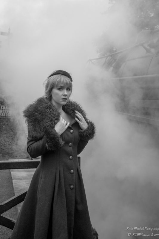 a woman in a coat and hat standing in front of a steam train