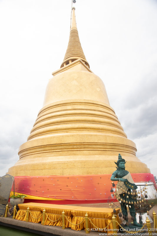 a large gold pagoda with a statue of a man in a green robe with Wat Saket in the background