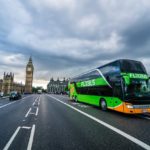 a green and orange bus on a road with a clock tower in the background