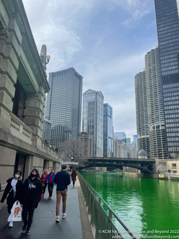 a group of people walking on a sidewalk next to a body of water with Chicago River in the background