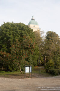 a sign in a park with trees and a building in the background