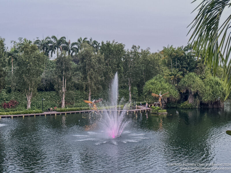 a water fountain in a pond with trees and a walkway